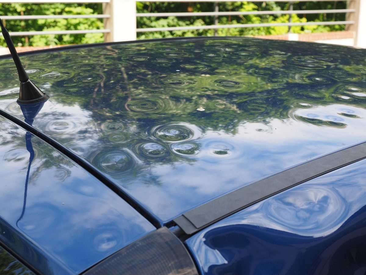 Voiture bleue avec des gouttes d'eau sur le toit, une antenne visible, ciel nuageux, végétation verte